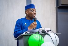Man in a blue traditional outfit and cap speaks into a microphone at a lectern with papers, green and white balloons nearby.