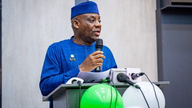 Man in a blue traditional outfit and cap speaks into a microphone at a lectern with papers, green and white balloons nearby.