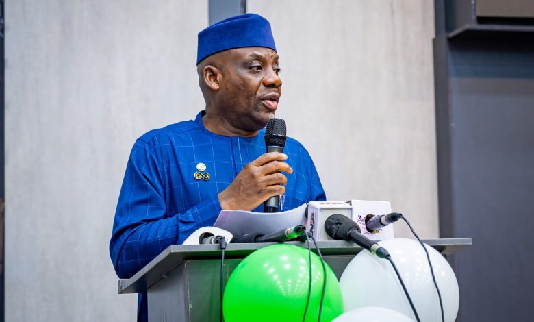 Tunji Alausa 1 - The Times of Abuja Man in a blue traditional outfit and cap speaks into a microphone at a lectern with papers, green and white balloons nearby.