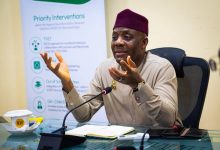 Man in beige traditional attire and maroon cap speaks into a microphone at a conference table, gesturing with his hands, notes open in front, banner behind him reading 'Priority Interventions'.