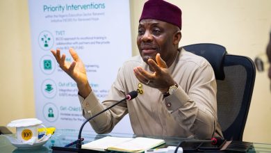 Man in beige traditional attire and maroon cap speaks into a microphone at a conference table, gesturing with his hands, notes open in front, banner behind him reading 'Priority Interventions'.