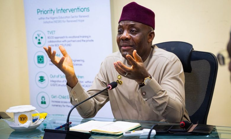 Tunji Alausa 2 - The Times of Abuja Man in beige traditional attire and maroon cap speaks into a microphone at a conference table, gesturing with his hands, notes open in front, banner behind him reading 'Priority Interventions'.