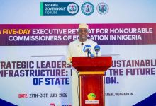 Man in white traditional attire speaks at a podium during a Nigeria Governors' Forum event, with a large banner behind him.