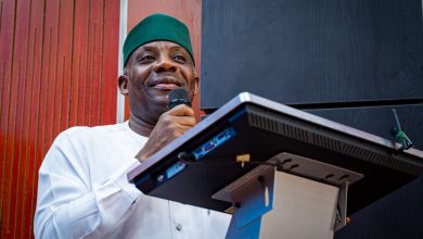 Man wearing a green hat and white traditional outfit speaks into a handheld microphone at a podium/lectern display setup behind him.