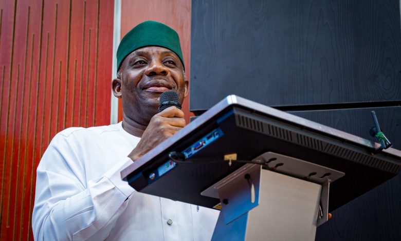Tunji Alausa - The Times of Abuja Man wearing a green hat and white traditional outfit speaks into a handheld microphone at a podium/lectern display setup behind him.