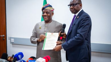 Two men exchange a certificate during a press briefing, with multiple microphones in front and a flag in the background.