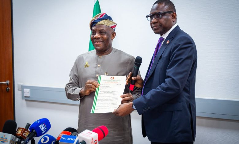 Two men exchange a certificate during a press briefing, with multiple microphones in front and a flag in the background.