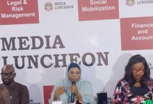 Woman in a light blue hijab speaks into a microphone at a media luncheon panel, with a 'Media Luncheon' backdrop.