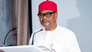 Man in white traditional attire and red cap speaking at a podium with a microphone.