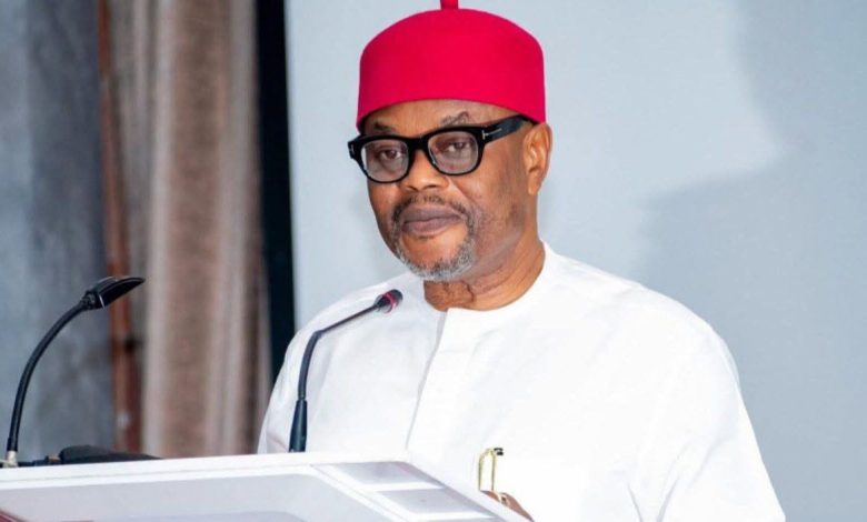 Uche Nnaji - The Times of Abuja Man in white traditional attire and red cap speaking at a podium with a microphone.