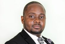 Professional headshot of a man in a dark suit and plaid gray tie, facing the camera with a neutral expression.
