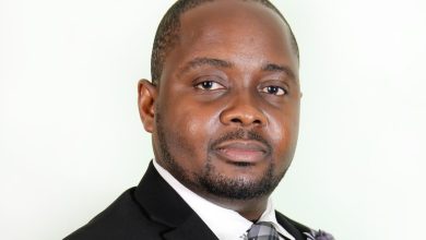 Professional headshot of a man in a dark suit and plaid gray tie, facing the camera with a neutral expression.