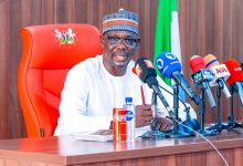 A man in white traditional attire and patterned cap speaks at a press conference, seated behind a row of microphones on a wooden table with a red high-back chair and Nigerian flag in the background.