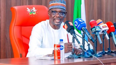 A man in white traditional attire and patterned cap speaks at a press conference, seated behind a row of microphones on a wooden table with a red high-back chair and Nigerian flag in the background.