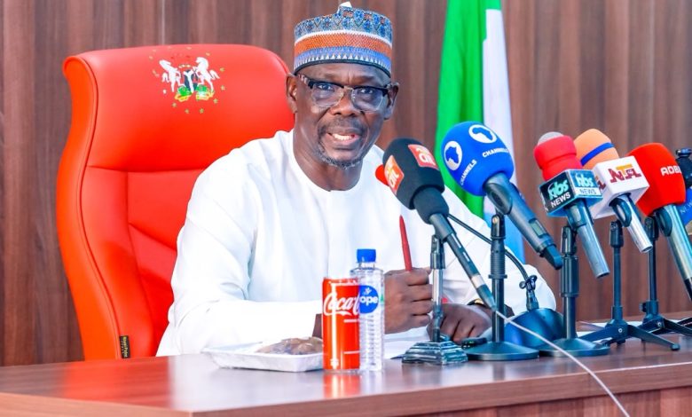 Abdullahi Sule - The Times of Abuja A man in white traditional attire and patterned cap speaks at a press conference, seated behind a row of microphones on a wooden table with a red high-back chair and Nigerian flag in the background.