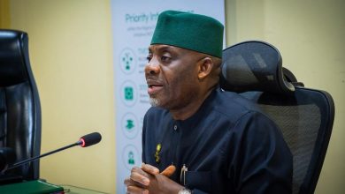 Man in a dark traditional outfit and green cap sits at a desk, hands clasped, speaking into a microphone in a conference room.
