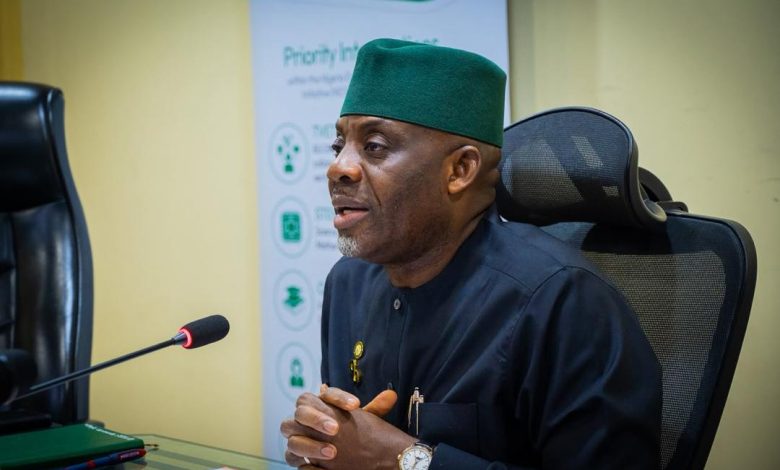 Alausa new - The Times of Abuja Man in a dark traditional outfit and green cap sits at a desk, hands clasped, speaking into a microphone in a conference room.