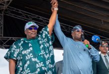 Two men on a stage raise their clasped hands in a show of victory; one wears a teal traditional outfit, the other a light blue shirt with a microphone in hand.
