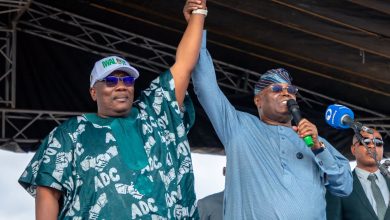 Two men on a stage raise their clasped hands in a show of victory; one wears a teal traditional outfit, the other a light blue shirt with a microphone in hand.