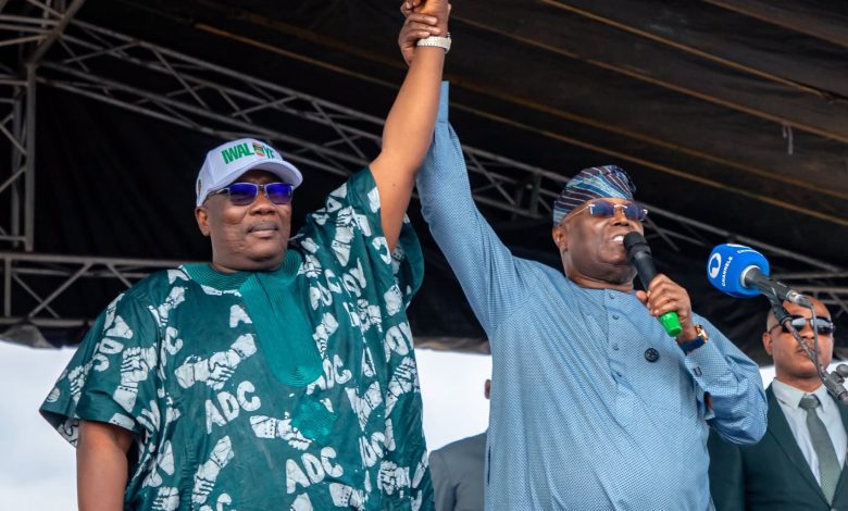 Two men on a stage raise their clasped hands in a show of victory; one wears a teal traditional outfit, the other a light blue shirt with a microphone in hand.