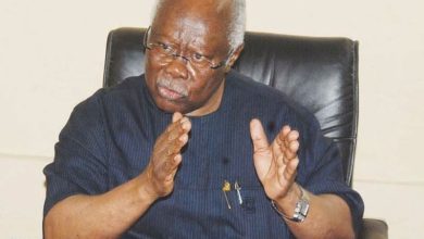 Older man with glasses in a dark blue shirt sits in a leather chair, gesturing with his hands while talking.