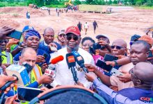 Man in white shirt and red cap speaks to a swarm of reporters holding microphones at a construction site.