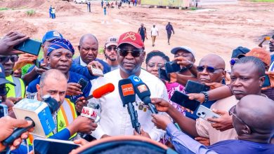 Man in white shirt and red cap speaks to a swarm of reporters holding microphones at a construction site.