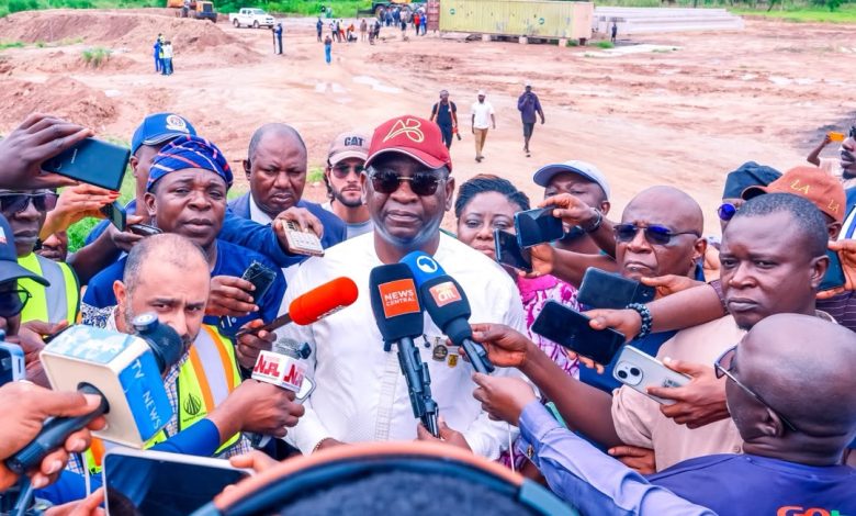 IMG-20260807-WA0018 - The Times of Abuja Man in white shirt and red cap speaks to a swarm of reporters holding microphones at a construction site.