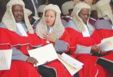 Group of judges wearing red robes and white wigs seated with documents at a formal ceremony.