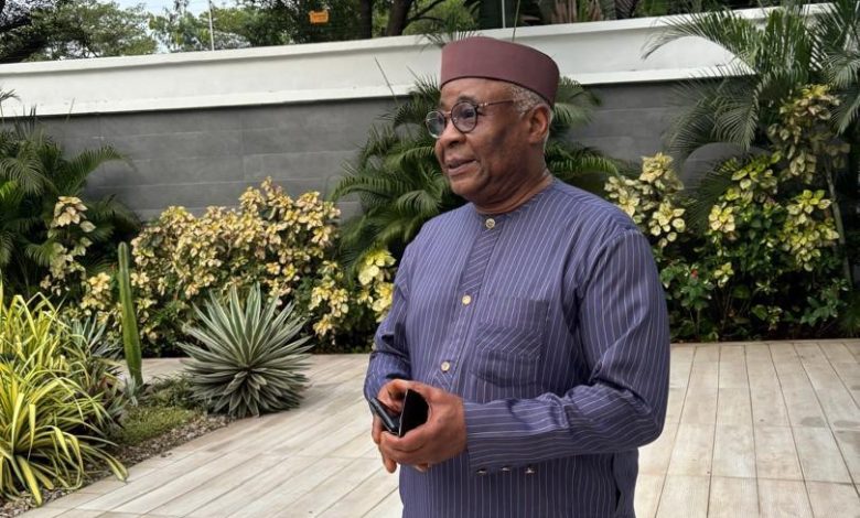 Older man wearing a maroon cap and striped navy tunic standing outdoors with a phone in his hands, looking to the side.