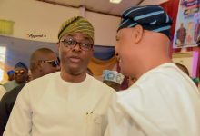 Man in white traditional attire and a striped cap talks with another man at an indoor social gathering, with guests in the background.