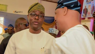 Man in white traditional attire and a striped cap talks with another man at an indoor social gathering, with guests in the background.