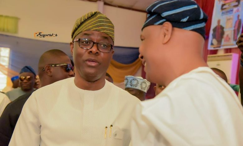Makinde and Wole Arisekola - The Times of Abuja Man in white traditional attire and a striped cap talks with another man at an indoor social gathering, with guests in the background.