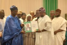 Group of dignitaries in traditional Nigerian attire at an award ceremony, exchanging a green certificate binder with a portrait on the page while others look on.