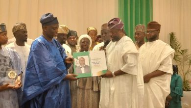 Group of dignitaries in traditional Nigerian attire at an award ceremony, exchanging a green certificate binder with a portrait on the page while others look on.