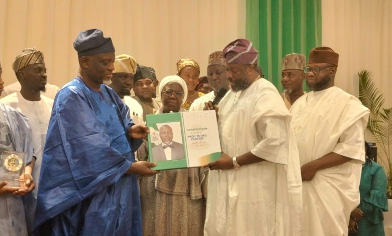 Oloyede and Aina - The Times of Abuja Group of dignitaries in traditional Nigerian attire at an award ceremony, exchanging a green certificate binder with a portrait on the page while others look on.