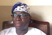 Portrait of a man wearing a blue and white patterned headwrap and glasses, seated in a brown leather chair in an indoor setting.