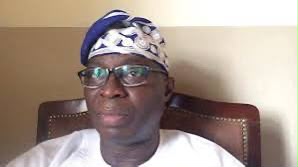 Portrait of a man wearing a blue and white patterned headwrap and glasses, seated in a brown leather chair in an indoor setting.
