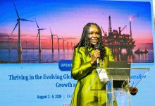 Female speaker in lime-green attire presenting at a conference, with wind turbines and an offshore oil-rig backdrop at sunset.