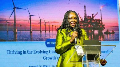 Female speaker in lime-green attire presenting at a conference, with wind turbines and an offshore oil-rig backdrop at sunset.