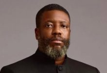 Portrait of a man with a gray beard wearing a black high-collar jacket, looking at the camera against a gray background.