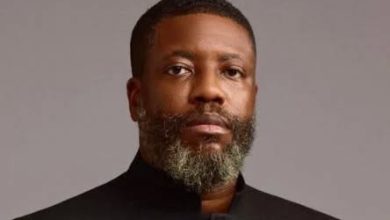 Portrait of a man with a gray beard wearing a black high-collar jacket, looking at the camera against a gray background.