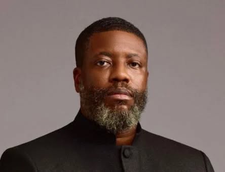 Segun Aina - The Times of Abuja Portrait of a man with a gray beard wearing a black high-collar jacket, looking at the camera against a gray background.