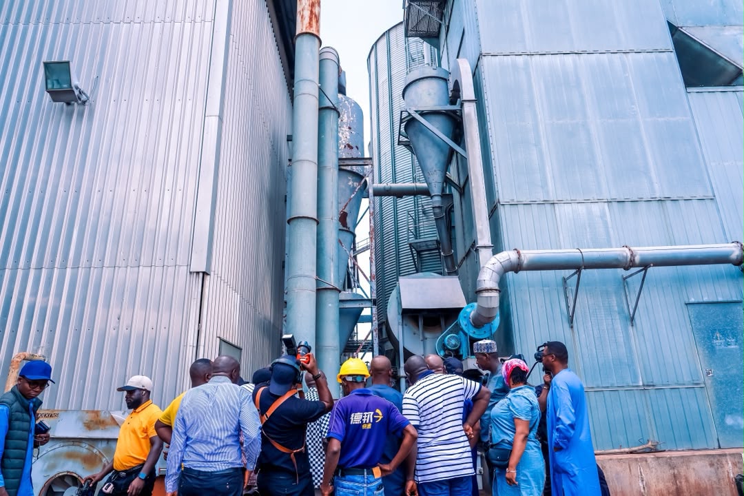 Taraku Mills 1 - The Times of Abuja Group of workers in hard hats and safety gear inspecting a large blue industrial facility with pipes and ducts between metal walls.