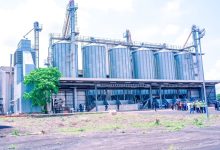 Industrial facility with tall metal silos and a two-story structure; workers in high-visibility vests stand outside on a sunny day
